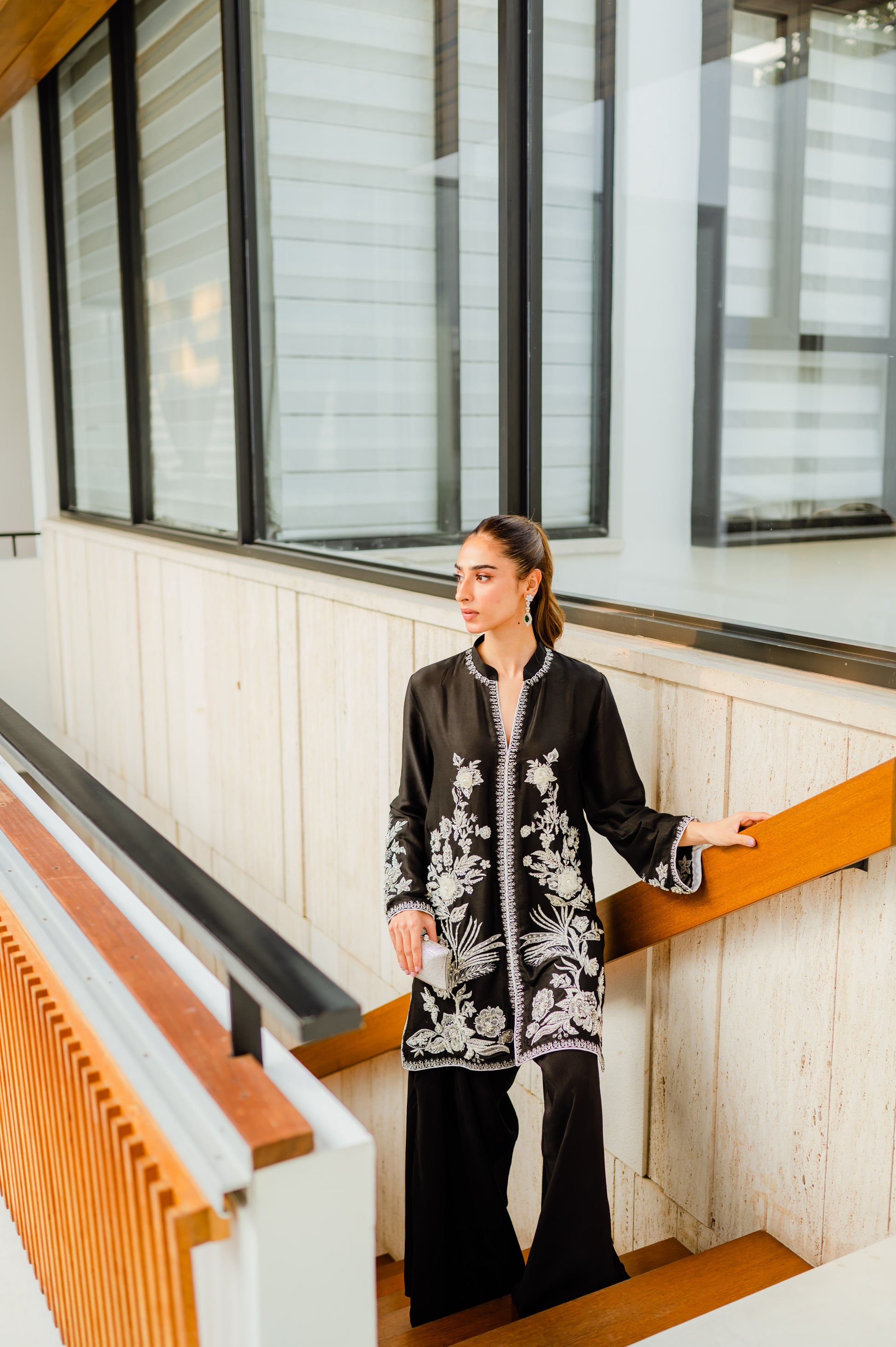 Woman wearing a hand-worked black jacket with Parsi pants, ideal for party and wedding wear, standing on stairs indoors.