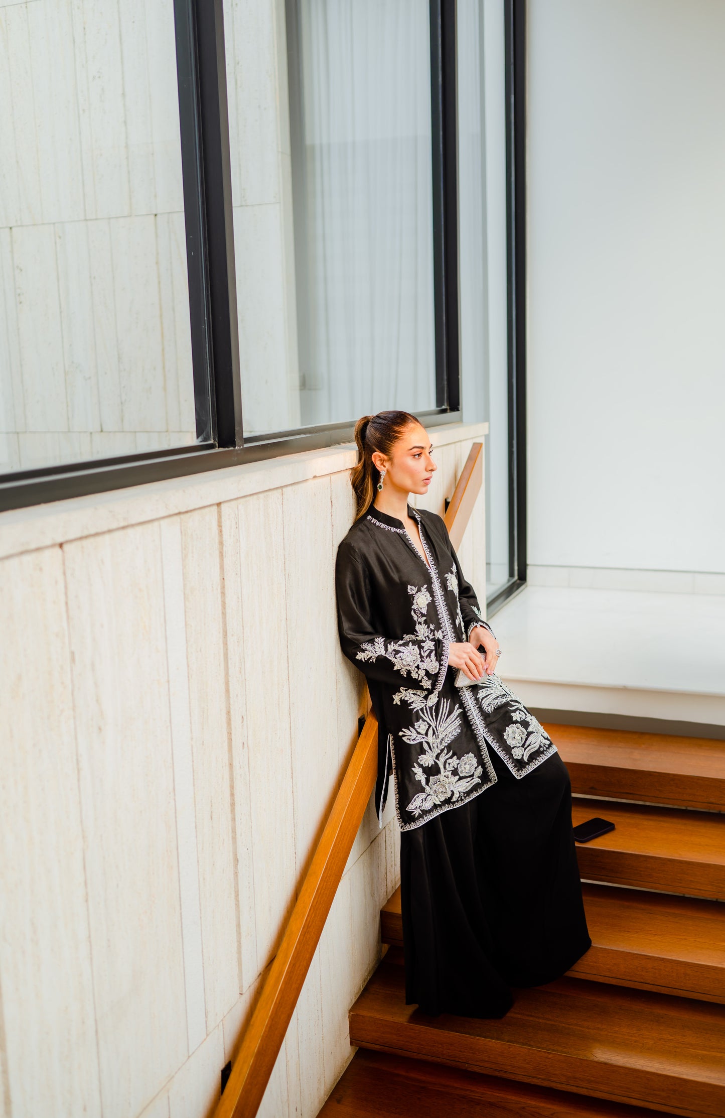 Woman wearing hand worked black jacket with Parsi pants, sitting on stairs in elegant setting, perfect for party or wedding wear.